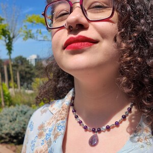 Bright sunlight shows the stone clearly — round violet beads of slightly varying depth, silver-tone diamond spacers set between them, and a pale lilac teardrop pendant crossed by one deeper purple band.