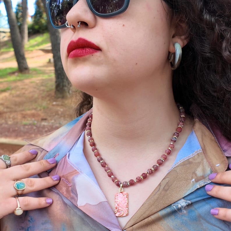 Close daylight view of the strand at the base of the neck: brick-red crackle beads, sparkling clear rhinestone rondelles, and the coral pendant with its raised gold lines resting against a pastel shirt.