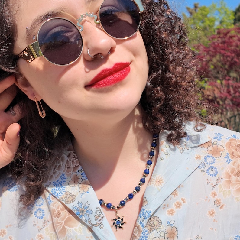 Sunlit outdoor shot from slightly above: the strand of deep blue beads runs from under the hair down the side of the neck to the gold sun pendant resting low against a pale floral shirt.