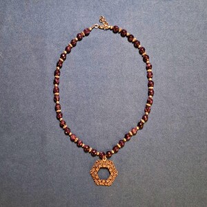 Deep purple faceted round beads laid in an oval on blue-grey fabric, a small metal spacer between each one, with an openwork hexagon pendant of tiny flowers at the bottom and a clasp and extender chain at the top.