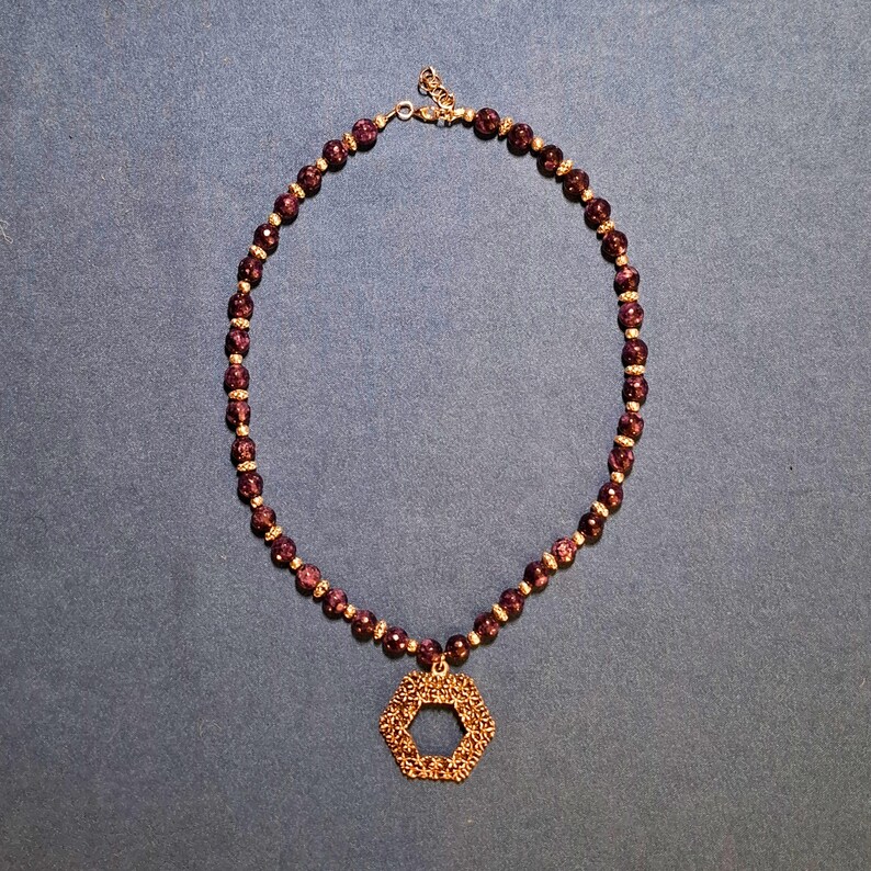 Deep purple faceted round beads laid in an oval on blue-grey fabric, a small metal spacer between each one, with an openwork hexagon pendant of tiny flowers at the bottom and a clasp and extender chain at the top.
