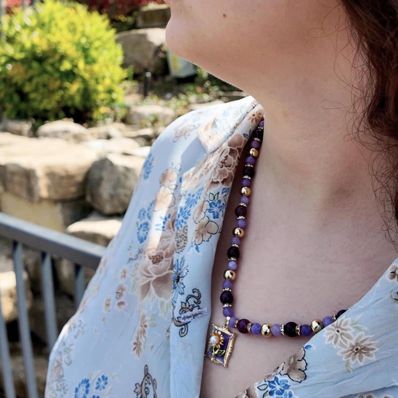Bright sun on the strand, showing the polished surface of the round beads and the tiny clear stones in the rondelles, with the gold-framed lilac enamel pendant angled against the blouse.