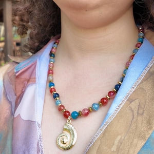 Close view of the strand worn at the base of the throat, red and blue crackled beads alternating with small gold spacers, the ribbed gold spiral pendant with its cream shell centre resting below.