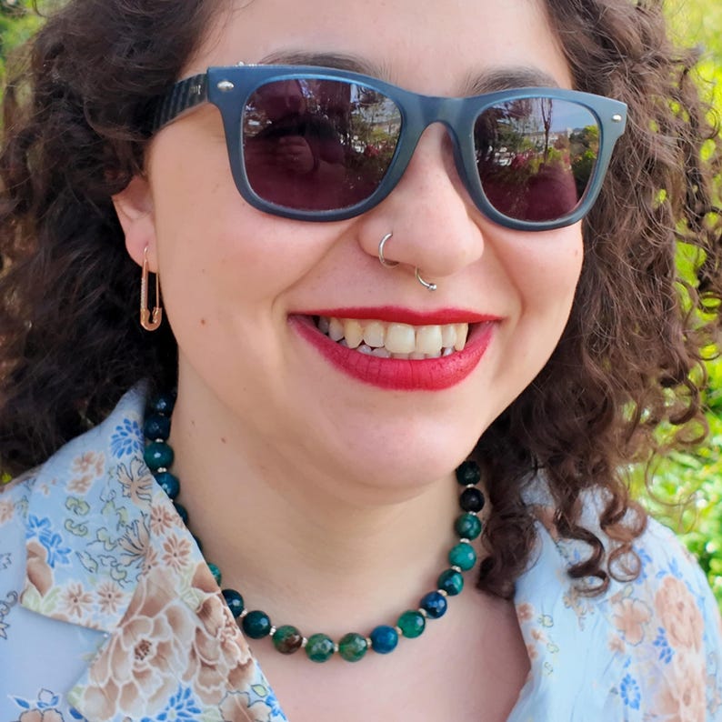 Close daylight angle on the necklace curving along the collarbone, deep green and blue faceted beads alternating, with one mottled brown-green bead sitting near the centre front.