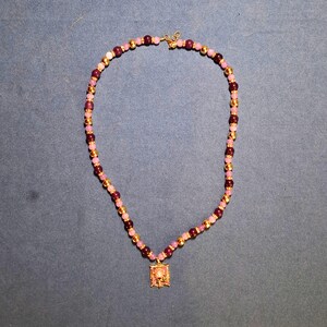 Beaded necklace laid in a loop on grey-blue cloth, alternating pale lilac and deep plum polished round beads with gold-tone balls and sparkling rondelles, a small rectangular enamel flower pendant at the front.