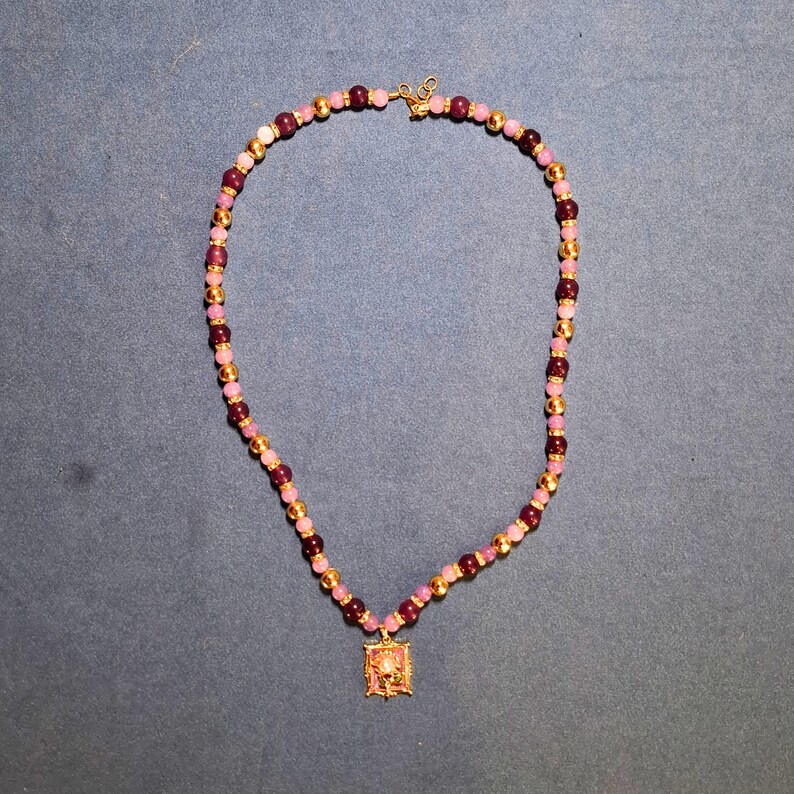 Beaded necklace laid in a loop on grey-blue cloth, alternating pale lilac and deep plum polished round beads with gold-tone balls and sparkling rondelles, a small rectangular enamel flower pendant at the front.