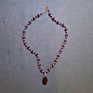 Deep blue beaded necklace laid in an open loop on grey-blue cloth, round stone beads alternating with sparkling rondelle spacers and tiny faceted beads, a smooth teardrop pendant hanging at the bottom and a clasp with extender at the top.