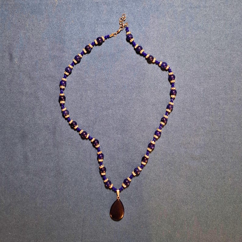Deep blue beaded necklace laid in an open loop on grey-blue cloth, round stone beads alternating with sparkling rondelle spacers and tiny faceted beads, a smooth teardrop pendant hanging at the bottom and a clasp with extender at the top.