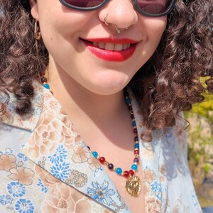 Close daylight view of the strand curving over the collarbone, showing the repeating red, purple and blue rounds with tiny gold spacers between them and the heart pendant at the front.