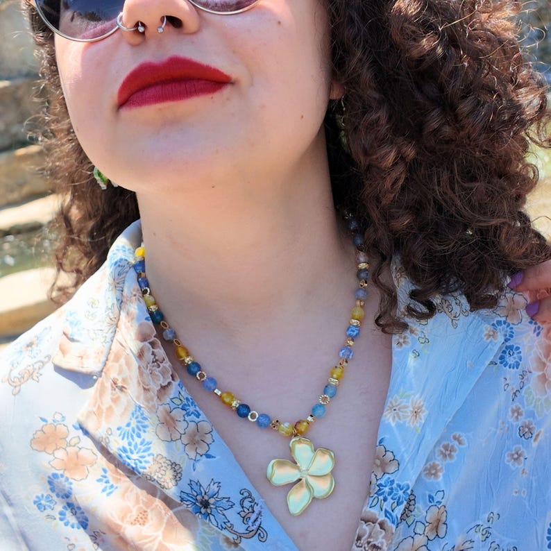 Close view of the necklace worn open over a blue floral shirt, the blue and mustard beads following the collarbone and the gold flower pendant resting at the top of the chest.
