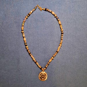 The whole necklace laid out in an oval on grey-blue cloth under warm indoor light, dark beads alternating with small gold rondelles and clear rhinestone rings, the round gold medallion resting at the bottom of the loop.