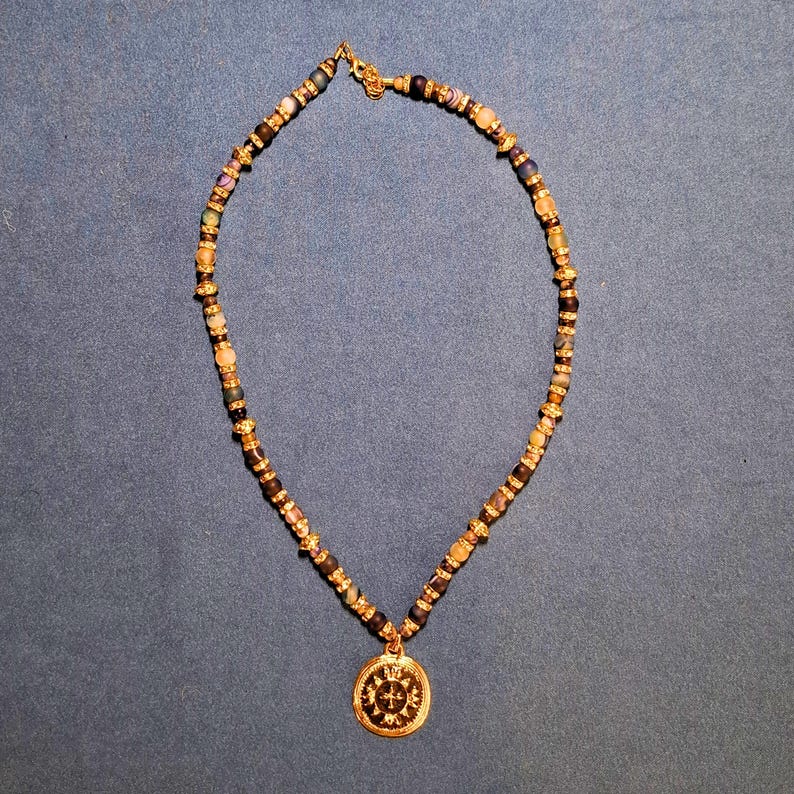 The whole necklace laid out in an oval on grey-blue cloth under warm indoor light, dark beads alternating with small gold rondelles and clear rhinestone rings, the round gold medallion resting at the bottom of the loop.