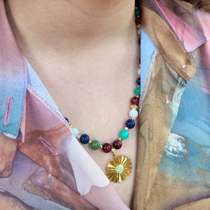 The necklace seen from slightly above, glossy round beads catching the light along the collarbone and the gold sunburst disc, about the width of three beads, hanging against the open patterned shirt.