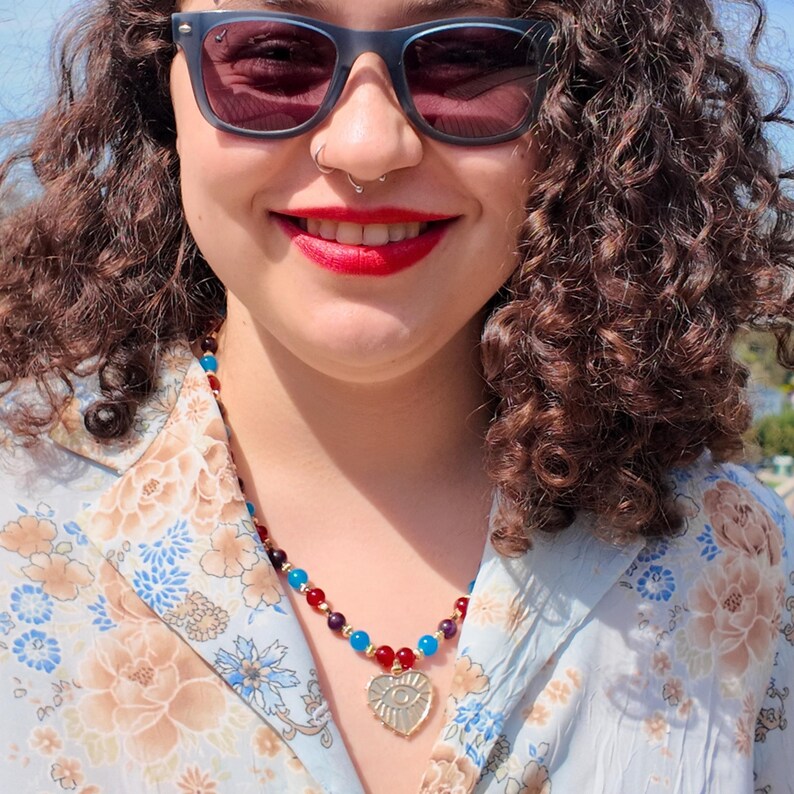 The necklace seen from above against a blue floral shirt, the small bright beads running down in a red, purple and blue sequence to the gold heart pendant with its engraved eye.