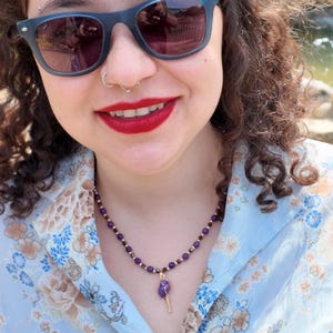 A smiling woman in a pale blue floral shirt wears the short purple bead strand at the base of her neck, the gold-set nugget pendant resting at the opening of her collar.