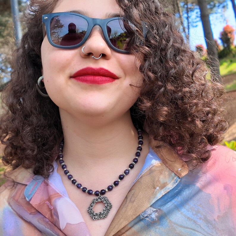 Seen from below with the head tilted back, the purple bead strand crosses the collarbones and the open hexagon pendant hangs free over the collar of a soft printed blouse.