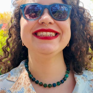 Front-on daylight view of the strand resting at the base of the neck, evenly sized faceted beads in teal, forest green and midnight blue separated by small metal spacers.