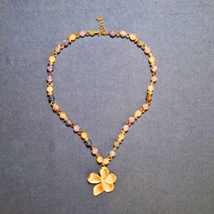 Beaded necklace laid flat on blue-grey cloth: round crackled beads in lilac-blue, honey and olive with gold-tone spacers, ending in a sculpted five-petal gold flower pendant at the centre front.