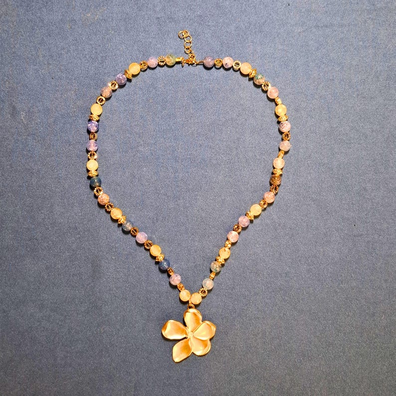 Beaded necklace laid flat on blue-grey cloth: round crackled beads in lilac-blue, honey and olive with gold-tone spacers, ending in a sculpted five-petal gold flower pendant at the centre front.