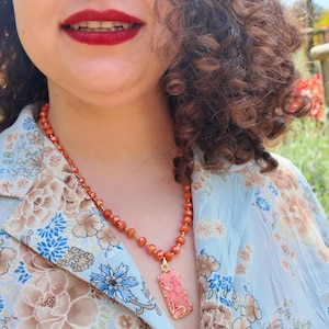 Puede incluir: Un collar con pequeñas cuentas redondas de tonos anaranjados ensartadas en una cadena dorada, terminado con un colgante rectangular. El colgante tiene un patrón texturizado de remolinos rosas y naranjas en un marco de metal dorado.