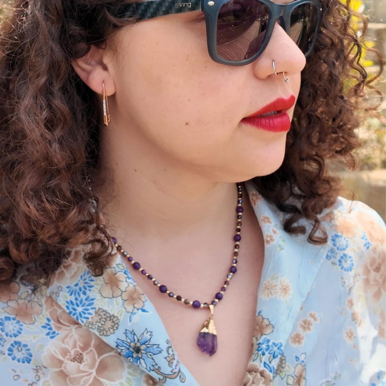 Front view outdoors: the strand of violet beads and gold-tone spacers sits on the collarbone, with the gold-capped purple crystal pendant dropping just into the open neckline.