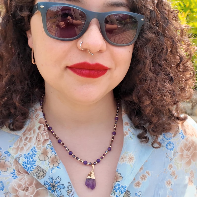 Close view from above of the necklace worn over a pale blue floral shirt, the purple and bronze beads following the base of the neck and the gold-capped violet crystal drop resting on the chest.