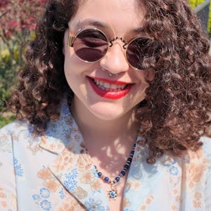 Worn in bright sun under an open floral blouse, the royal-blue beads sit at the base of the throat and the small gold sun pendant hangs in the V of the neckline, dappled shade falling across the skin.