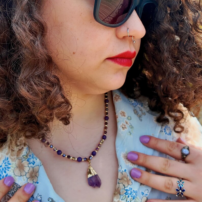 Worn in sunlight beside the collar of a blue and beige floral shirt, the small purple beads catch the light and the rough amethyst-coloured pendant hangs in its hammered gold cap.