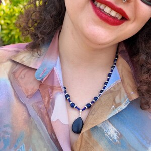 Outdoor portrait in a park showing the full necklace over a pastel watercolour shirt, the dark blue strand sitting around the base of the neck with the teardrop pendant hanging at the centre for scale.