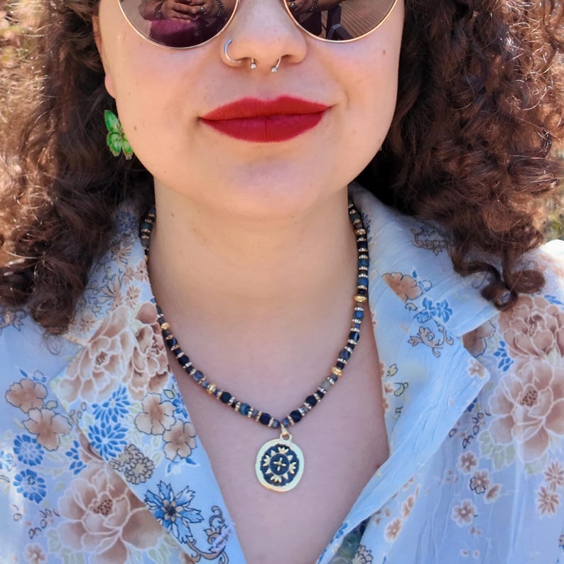 Outdoor three-quarter view in bright sun, the navy beaded strand curving across the collarbone with gold spacers catching the light and the round medallion low on the chest.