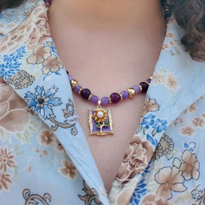 The necklace worn over an open pale blue floral blouse, lilac and plum beads curving under the collar and the gold-framed violet enamel pendant resting just below the collarbone.
