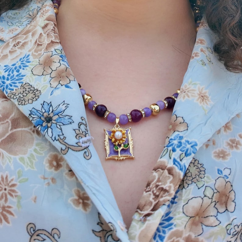 The necklace worn over an open pale blue floral blouse, lilac and plum beads curving under the collar and the gold-framed violet enamel pendant resting just below the collarbone.