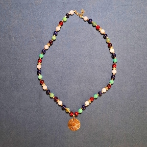 Beaded necklace laid in an oval on blue-grey fabric, round 8 mm beads in navy, cream, mint, teal and wine red with gold-tone spacers, a gold-tone lobster clasp at the top and a ridged gold sunburst disc hanging at the bottom.