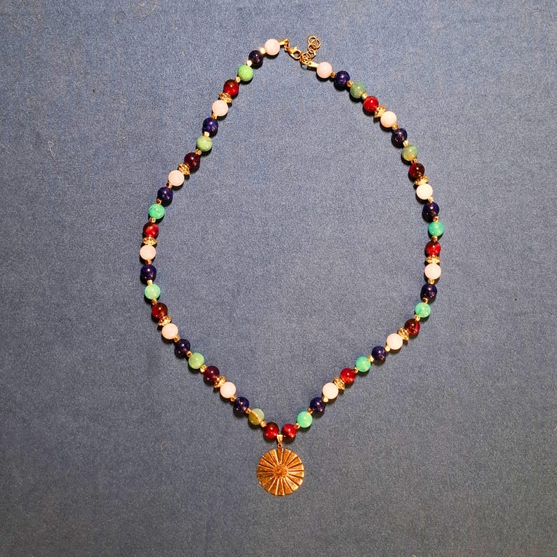 Beaded necklace laid in an oval on blue-grey fabric, round 8 mm beads in navy, cream, mint, teal and wine red with gold-tone spacers, a gold-tone lobster clasp at the top and a ridged gold sunburst disc hanging at the bottom.