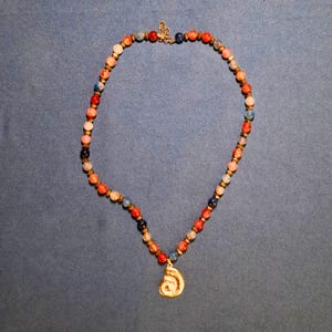 A single strand of 8 mm round beads in coral, red, blue, navy and pale grey, spaced with tiny gold-tone faceted beads, laid in an oval on grey-blue cloth with a gold spiral shell pendant at the bottom.