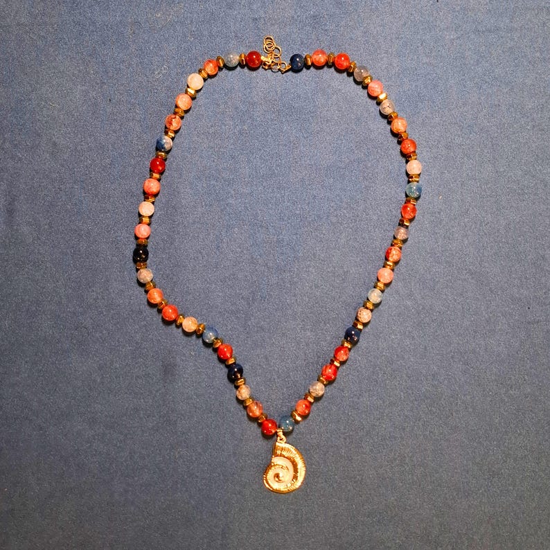 A single strand of 8 mm round beads in coral, red, blue, navy and pale grey, spaced with tiny gold-tone faceted beads, laid in an oval on grey-blue cloth with a gold spiral shell pendant at the bottom.