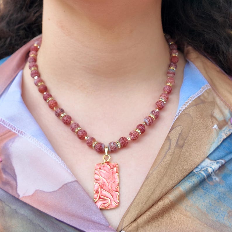 Outdoor shot of the necklace worn open-collar, hands resting near the neckline with stone rings, showing the strand's scale against the collar and the rectangular pendant dropping just below the collarbone.