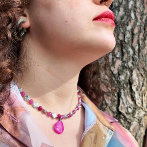 Close view of the wearer smiling in daylight, the pink bead strand sitting snug at the base of the neck with the glossy teardrop hanging centred below it.