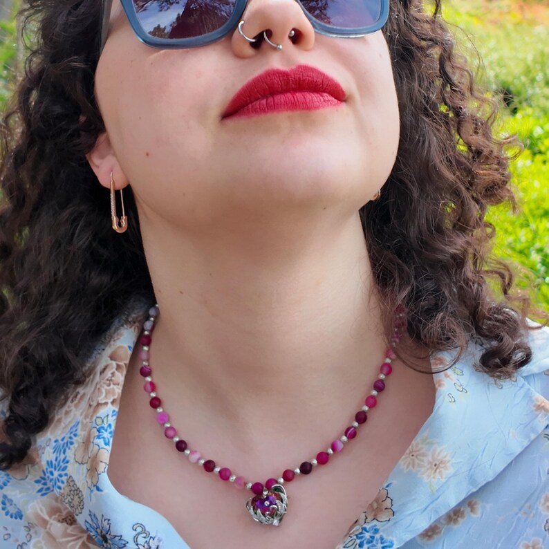 Worn outdoors over an open blue floral shirt, the strand of pink and burgundy beads follows the base of the neck and the heart pendant rests just below the hollow of the throat.