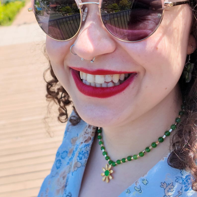 Worn in sunlight over a pale floral shirt, the green beaded strand sits at the base of the neck and the daisy pendant with its green centre rests just below the throat.