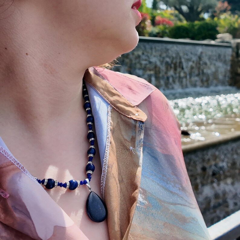 Front view of the necklace worn open-collar, deep blue round beads separated by bright rhinestone spacers, the smooth navy teardrop pendant sitting just below the collarbone against bare skin.