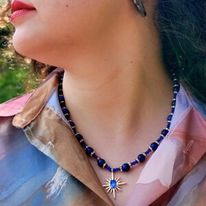 Close-up of the front of the necklace showing polished navy beads about 8 mm across, clear crystal rondelles in gold-tone settings, and the gold starburst pendant with its faceted blue stone.