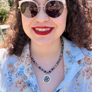 The necklace worn in daylight over an open blue floral shirt, navy beads with gold and rhinestone spacers sitting at the base of the neck, the gold medallion hanging just below the collarbone.