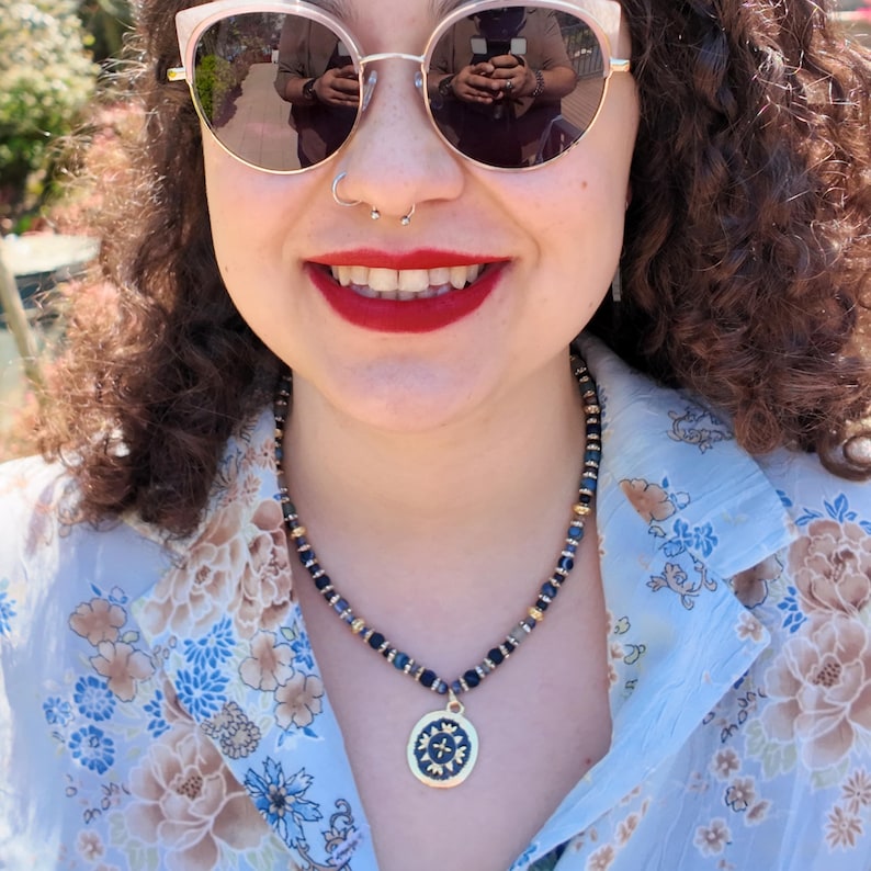 The necklace worn in daylight over an open blue floral shirt, navy beads with gold and rhinestone spacers sitting at the base of the neck, the gold medallion hanging just below the collarbone.