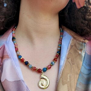 Sunlit angled view along the collarbone showing the white webbing inside the red and blue beads, the faceted gold spacers between them, and the gold spiral shell pendant below.