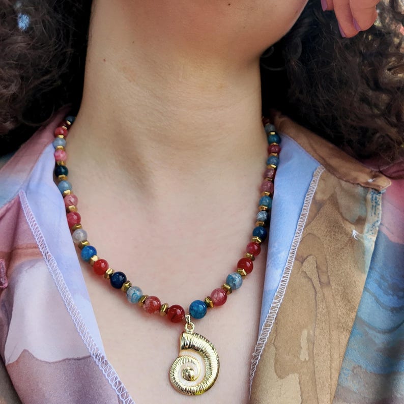 Sunlit angled view along the collarbone showing the white webbing inside the red and blue beads, the faceted gold spacers between them, and the gold spiral shell pendant below.