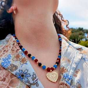 Sunlit shot of the necklace worn open-collar, the glossy red and blue beads catching light against the skin and the gold heart pendant hanging at the point of the shirt's V.