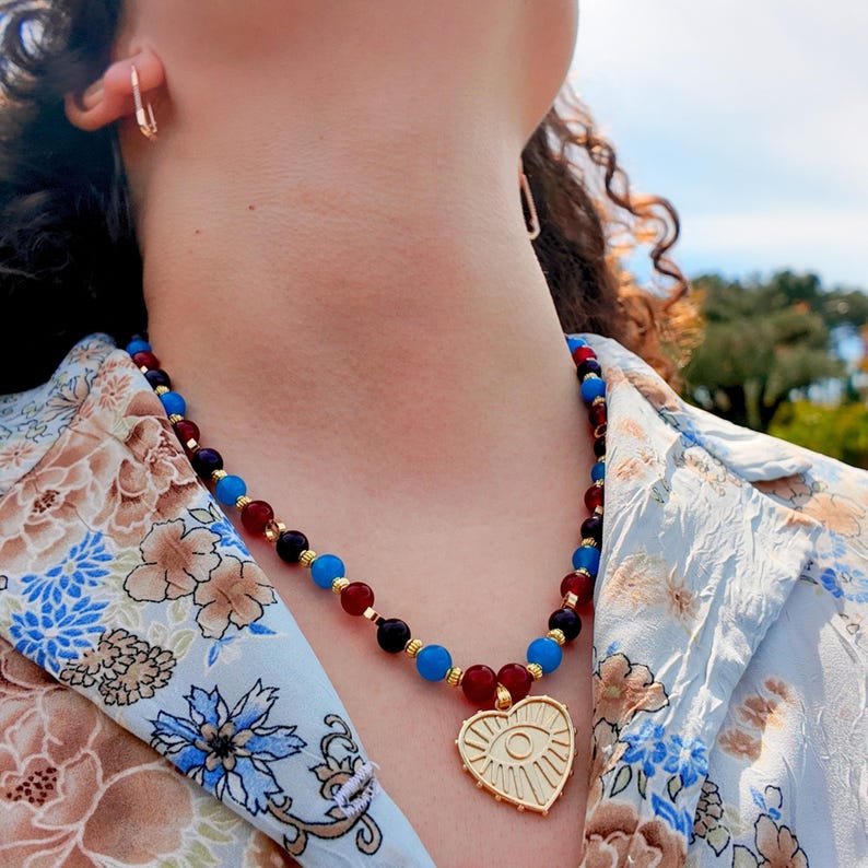 Sunlit shot of the necklace worn open-collar, the glossy red and blue beads catching light against the skin and the gold heart pendant hanging at the point of the shirt's V.