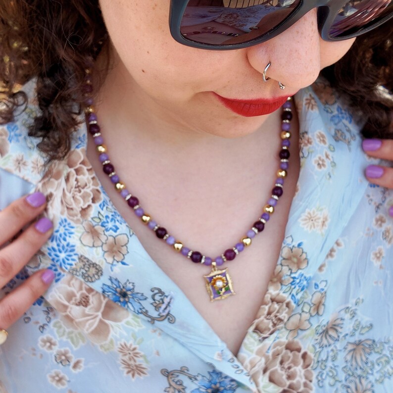 Side view of the strand passing over the shoulder and down the neck, small lilac beads and larger plum beads catching daylight, the rectangular enamel pendant sitting in the open collar.