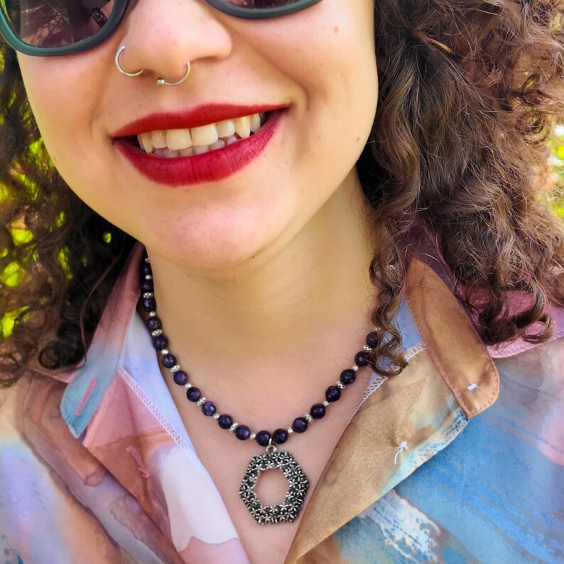 Side view in sunlight: the strand of small purple beads follows the side of the neck and the openwork hexagon pendant sits centred on the chest above an open collar.